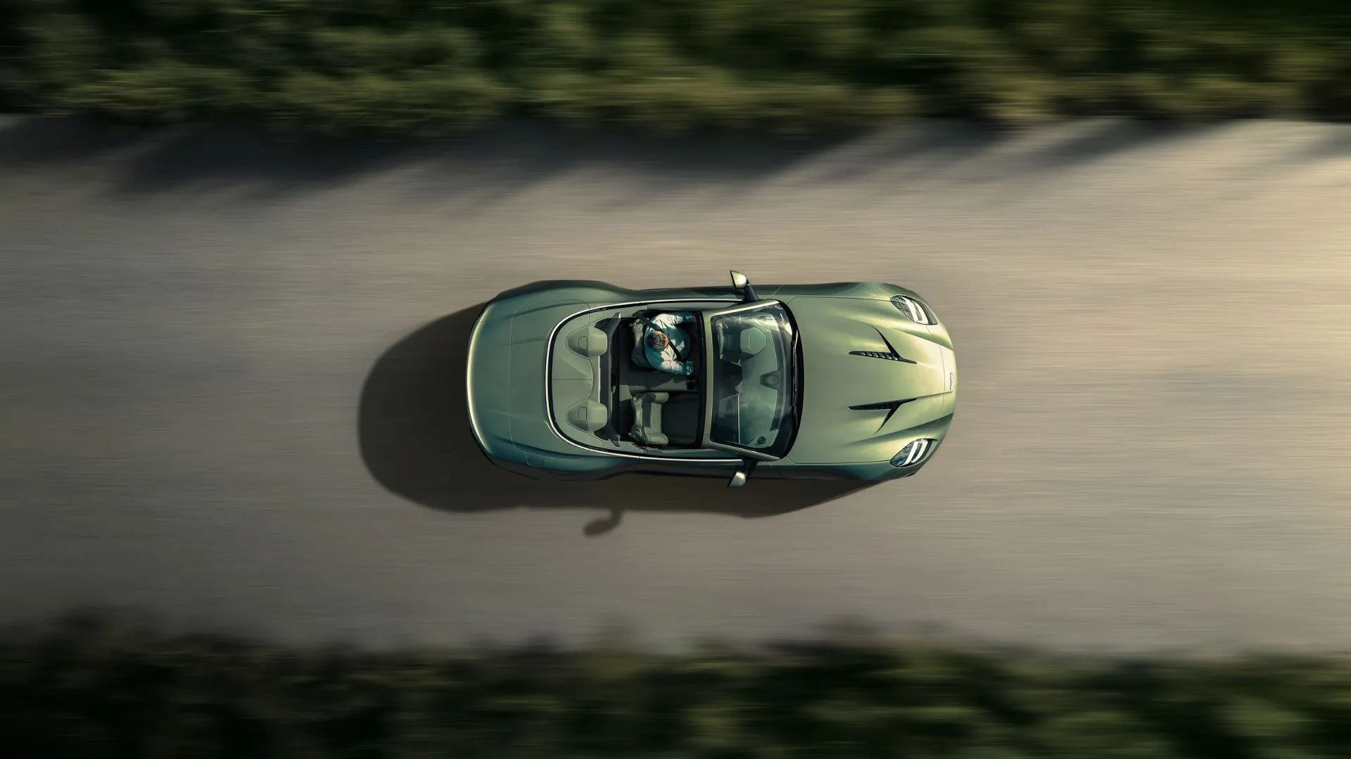 Aston Martin Vanquish Volante mui trần màu xanh ngọc, nhìn từ trên cao, đang lướt trên con đường nông thôn với cảnh sắc thiên nhiên tuyệt đẹp, tôn lên vẻ sang trọng và tự do.