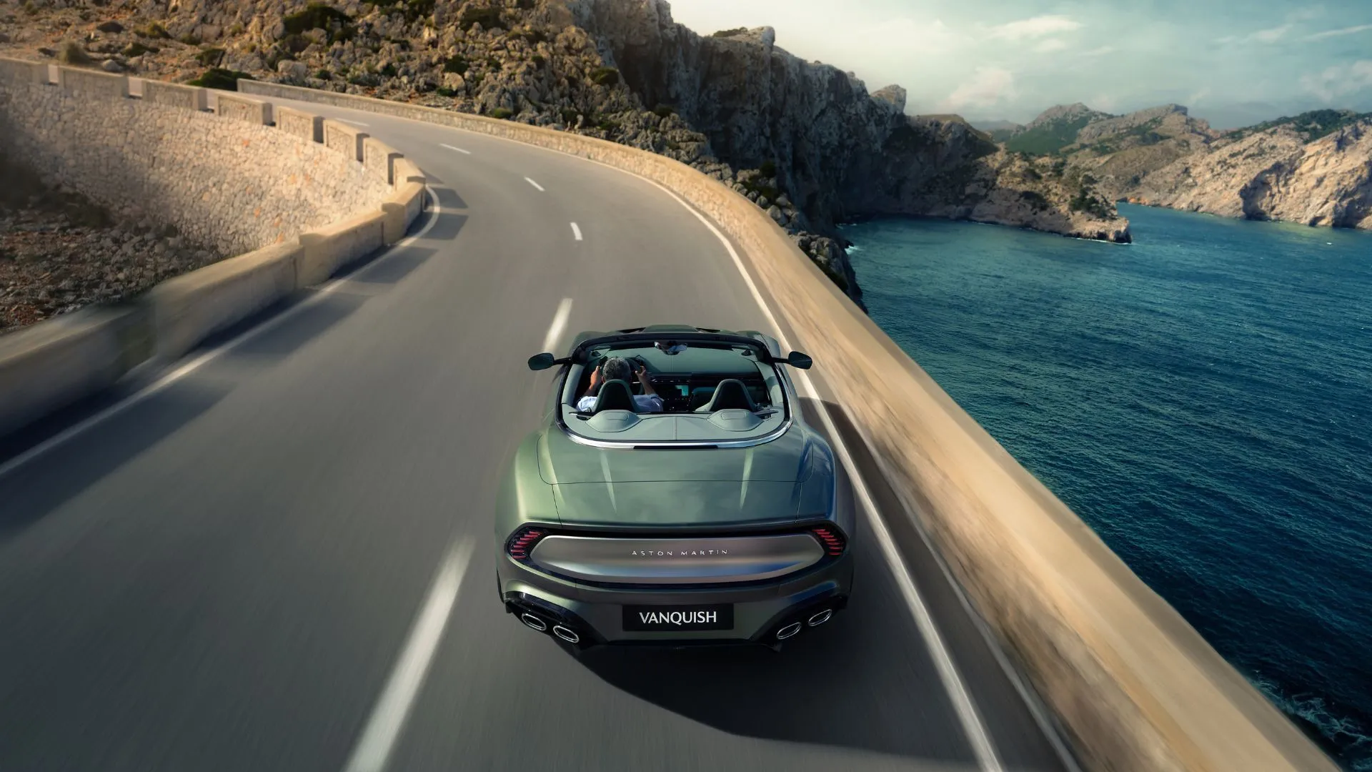 Aston Martin Vanquish Volante mui trần nhìn từ phía sau, đang di chuyển trên một con đường quanh co cạnh hồ nước, thể hiện sự mạnh mẽ và tinh tế của siêu xe Anh Quốc.