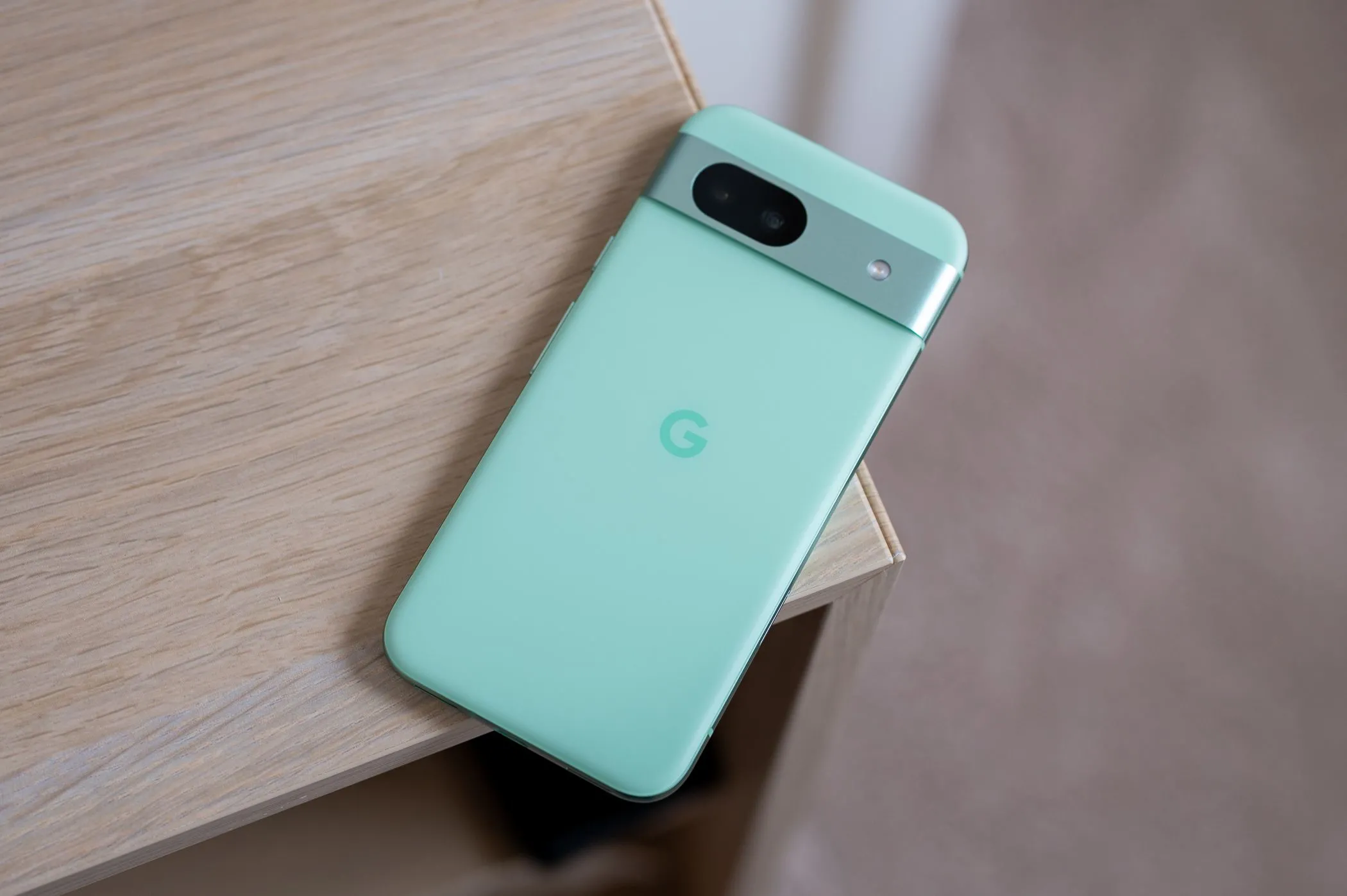 Điện thoại Pixel 8a màu xanh ngọc được đặt trên mép bàn, thể hiện thiết kế nhỏ gọn và viền màn hình đặc trưng.
